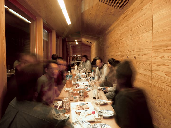 Group of travelers eating and talking around a long table inside a warm, wood-paneled Osaka lodging.
