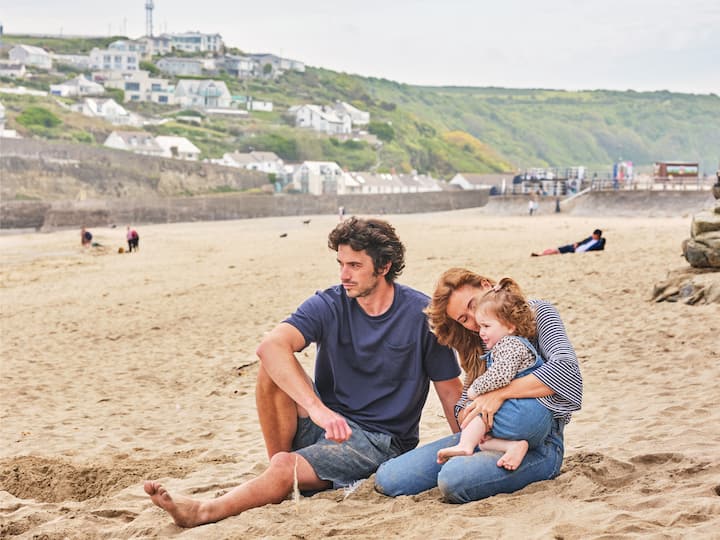 Family relaxing together on a sandy UK beach with a coastal village and green cliffs behind them.