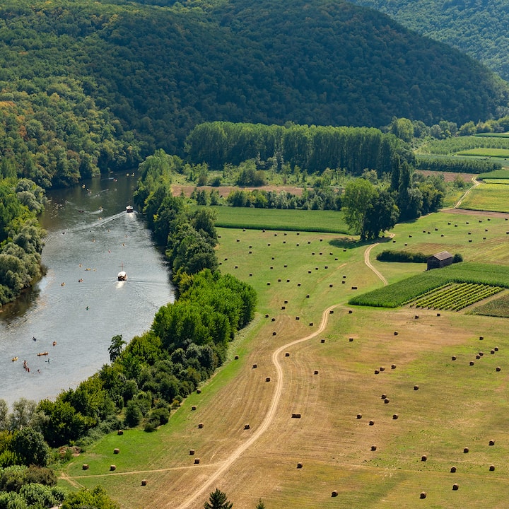 Aerial view of a winding river with kayaks and a boat, lush forest and hay-spotted fields.