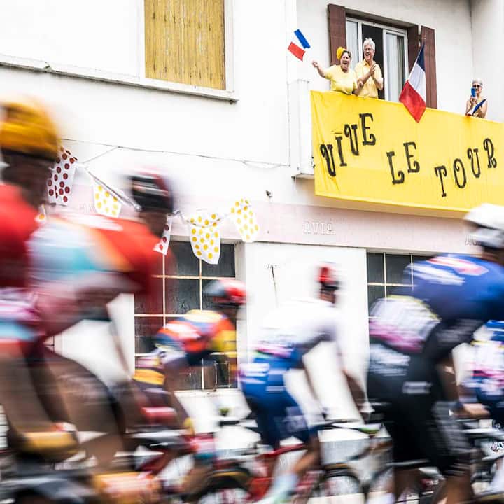 Blurred cyclists speed past a house as two fans wave French flags above a yellow “Vive le Tour” banner.