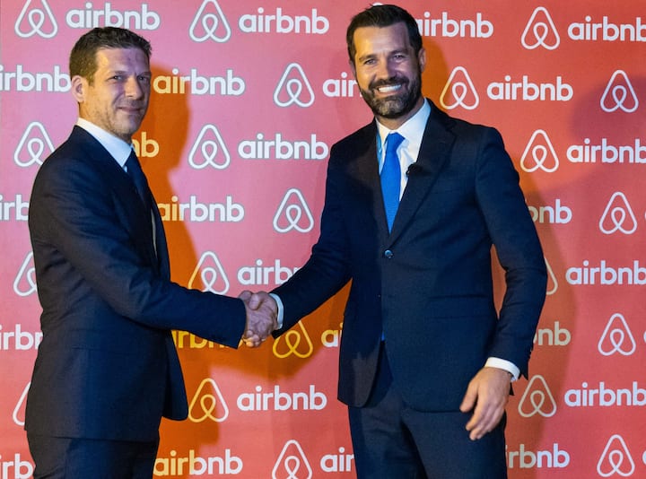 Two suited officials shake hands in front of an Airbnb step‑and‑repeat backdrop at a policy event.