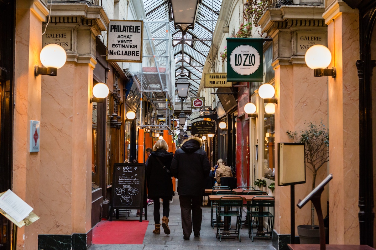 Two people stroll through a covered Paris passage lined with cafés, shops, and warm glowing lights.