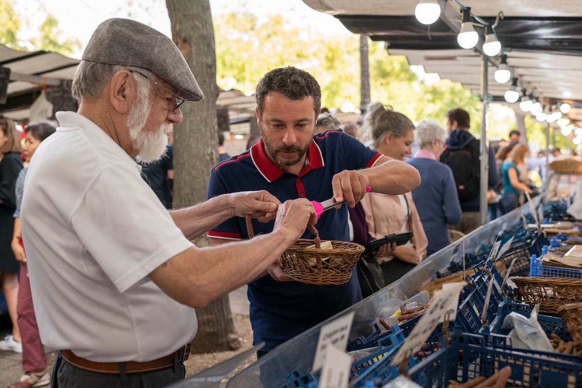 Travelers sampling local food at an outdoor market while browsing baskets of regional specialties.