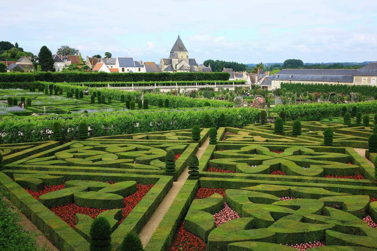 Ornate royal-style gardens outside Paris, with maze-like hedges and a village and church in the background.