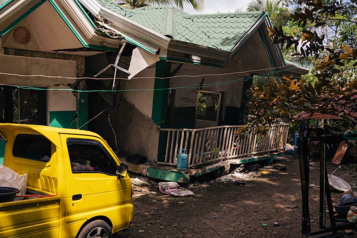 A home with significant structural damage including collapsed roof sections, cracked exterior walls, and a shifted foundation following an earthquake. A yellow vehicle is parked in the foreground amid scattered debris.