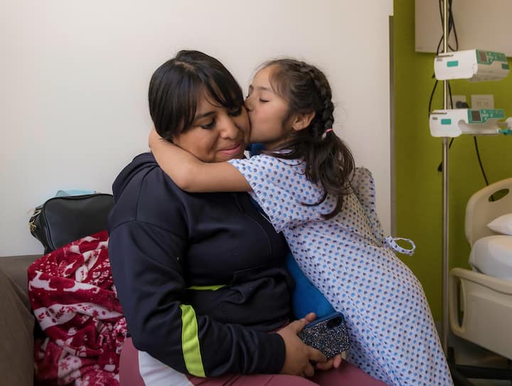 A young girl in a hospital gown kisses and embraces a woman seated beside her in a medical room, with an IV stand and hospital bed visible in the background.