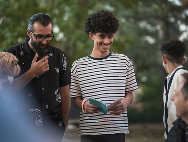 A young man in a striped t-shirt smiles while holding a small card or booklet, standing outdoors with a group of people and a dog partially visible in the background.