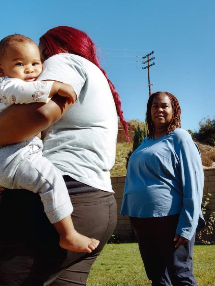 A woman with red hair holds a baby outdoors while another woman in a blue top stands nearby, with a clear blue sky and a grassy yard in the background.A woman with red hair holds a baby outdoors while another woman in a blue top stands nearby, with a clear blue sky and a grassy yard in the background.