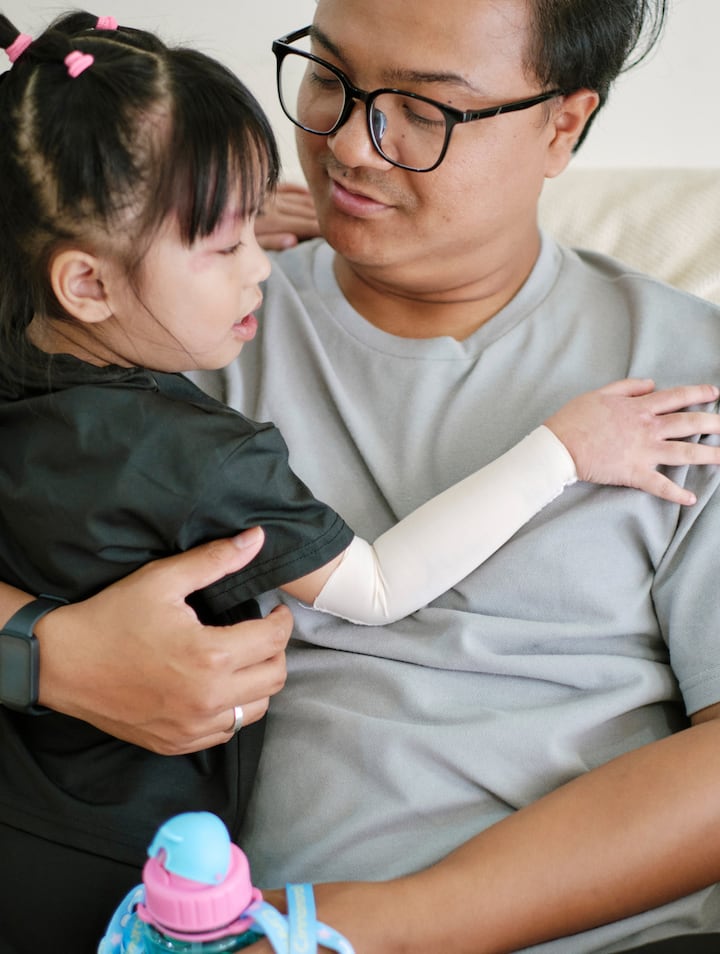 A man wearing glasses holds a young girl in his arms indoors, looking down at her warmly while she rests against him.A man wearing glasses holds a young girl in his arms indoors, looking down at her warmly while she rests against him.