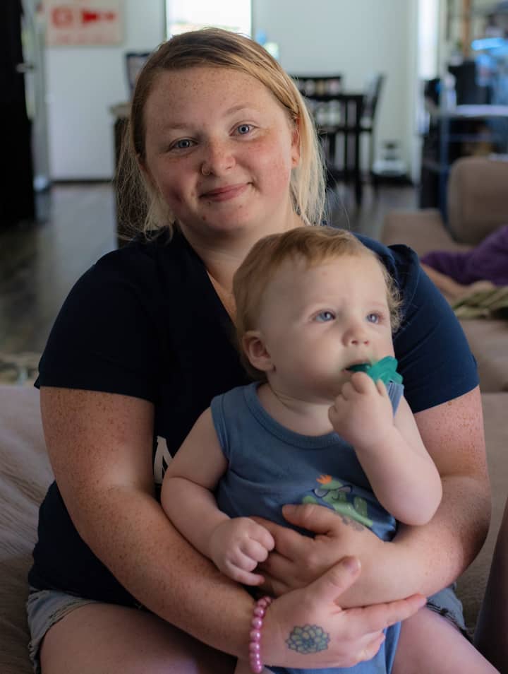 A young woman sits indoors holding a baby with a pacifier, smiling gently at the camera with a living space visible in the background.A young woman sits indoors holding a baby with a pacifier, smiling gently at the camera with a living space visible in the background.