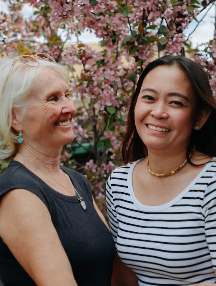 Two women smile together outdoors in front of a flowering tree with pink blossoms
