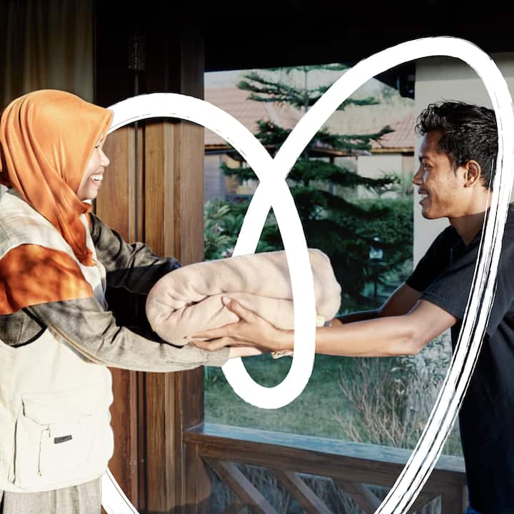 A woman in a hijab smiles while handing a folded blanket to a man at the entrance of a home, with a hand drawn heart  illustration overlaid across the image.
