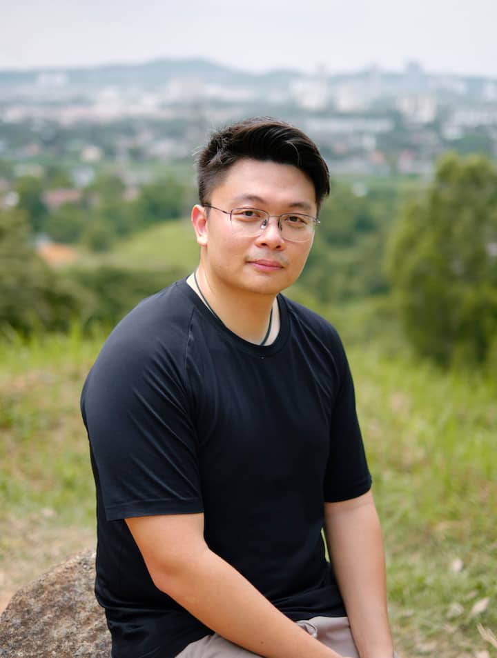 A man sitting outdoors on a hillside with a cityscape and greenery visible in the background.