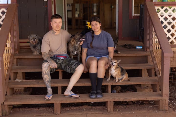 A family photo of Maile and Kai sitting on the front steps of their home with three dogs: Blackie, Tiny, and Noelle.