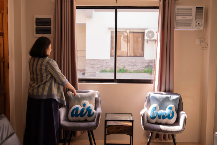 Host standing by a bright window in her living room, ready to welcome guests