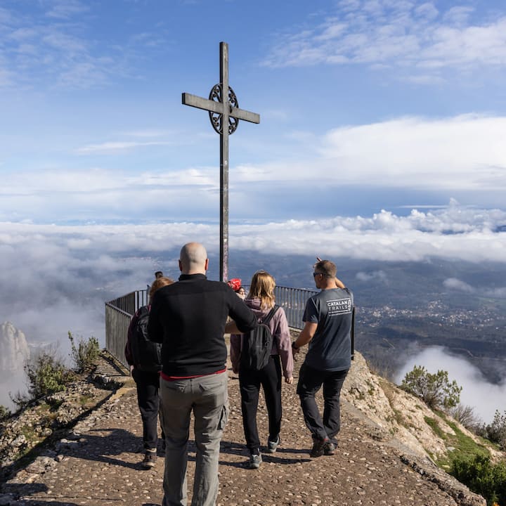 Varias personas caminan hacia un mirador de montaña en España. Están reunidas junto a una cruz alta sobre un valle lleno de nubes.