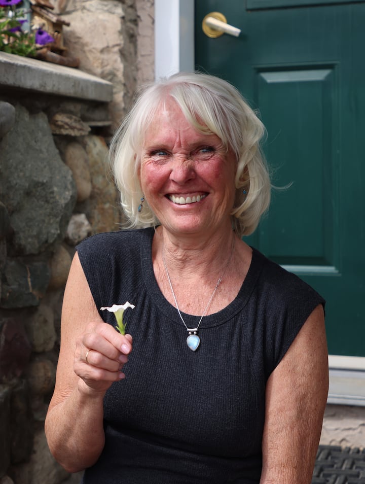 An older woman smiling in front of a stone wall with a green door, holding a small white flower.