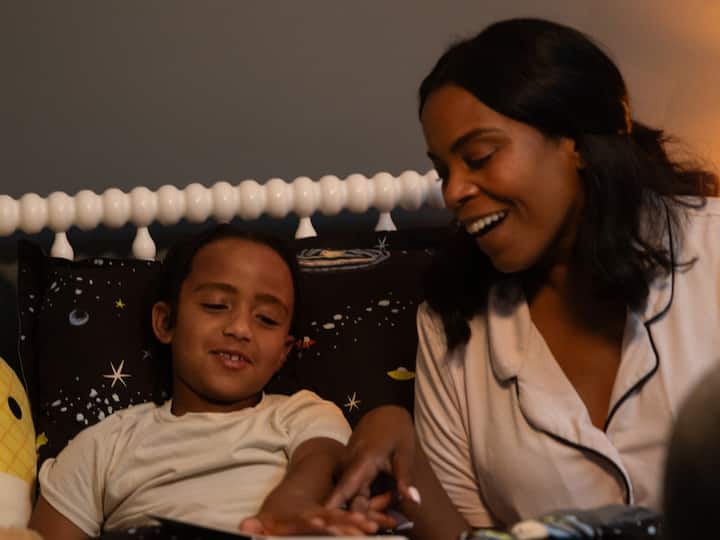 A woman smiles warmly at a young child lying in bed, in a softly lit bedroom.