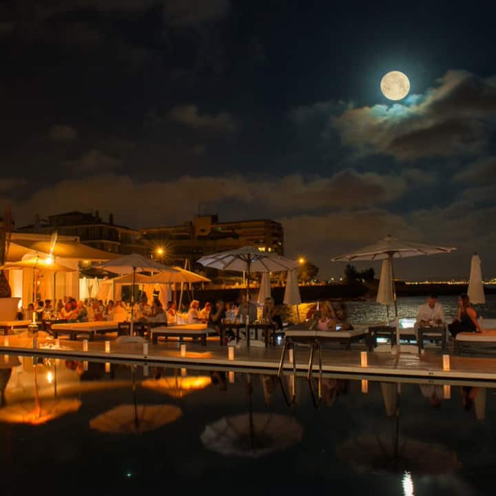 Varios viajeros se relajan en una terraza frente al mar en Mallorca bajo una brillante luna llena, listos para disfrutar de una experiencia para ver el eclipse.