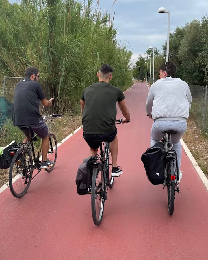 Unos ciclistas pedalean por un carril bici rojo a las afueras de Valencia, rodeados de hierba alta y árboles en un día nublado.