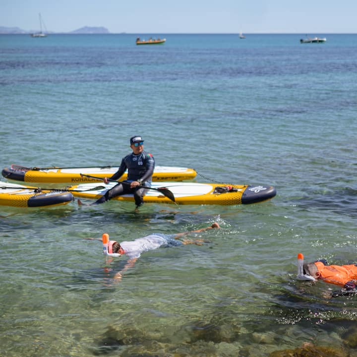 Varias personas practicando snorkel flotan cerca de un guía sentado en una tabla de paddle surf amarilla. Se encuentran junto a la costa de España desde la que se verá el eclipse.