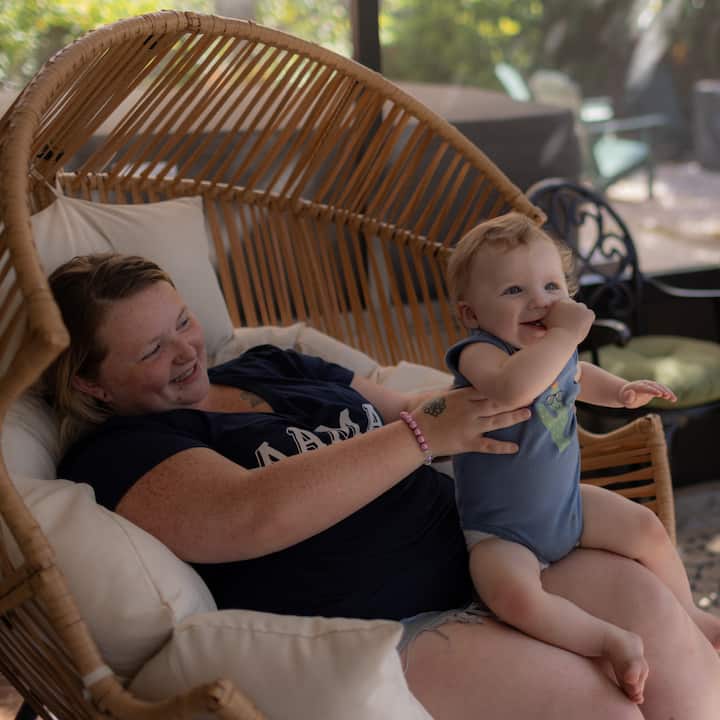 A woman laughs with a baby on her lap while seated in a rattan chair outdoors.