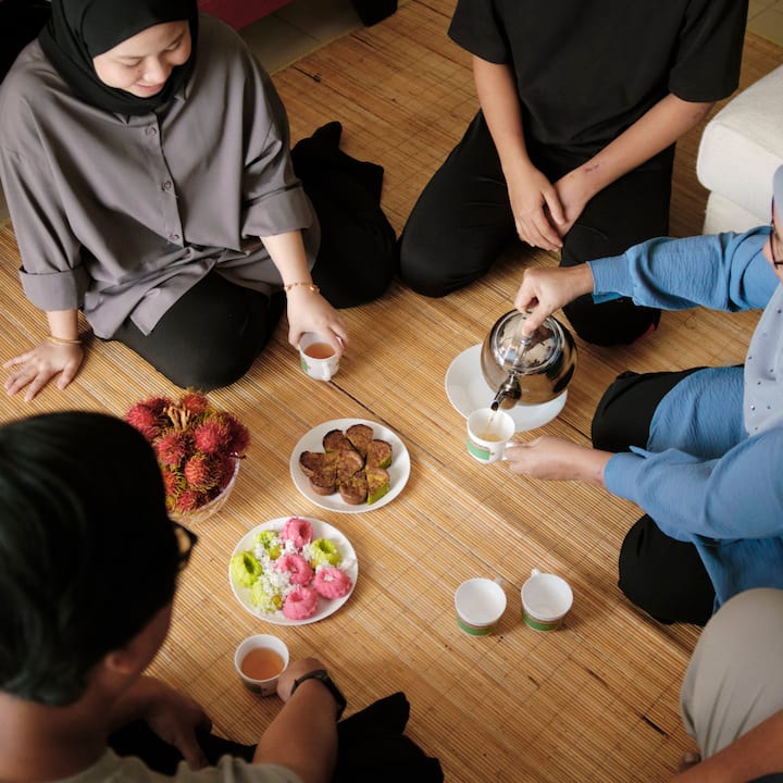 An overhead view of a group of people seated on a woven mat sharing tea and traditional snacks together.