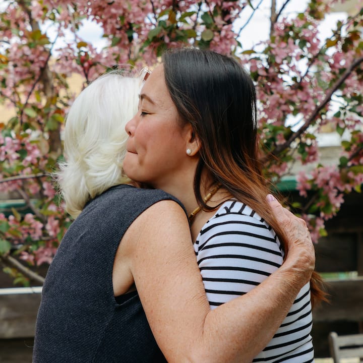 Two women embrace warmly outdoors in front of a flowering tree with pink blossoms.