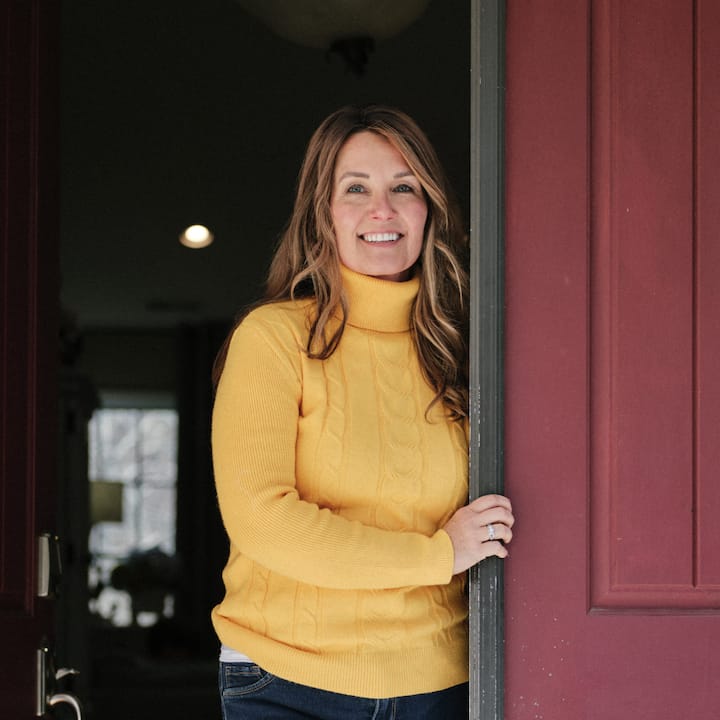 A woman smiles while standing in the open doorway of a home with a red door.