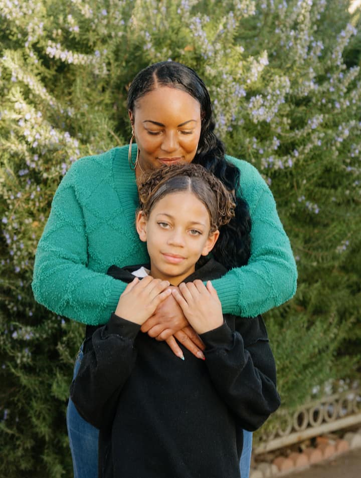 A mother stands behind her child outdoors, embracing them warmly in front of flowering greenery.