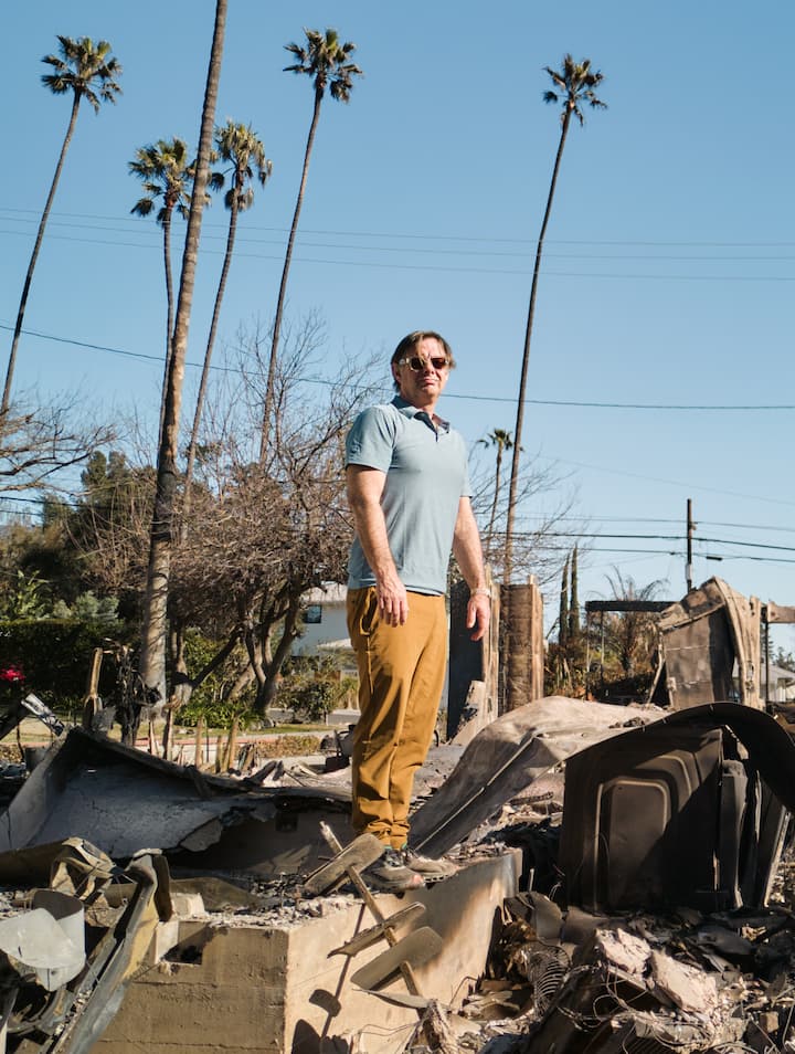 A man stands amid the charred rubble of a destroyed structure, with tall palm trees and a clear blue sky in the background.