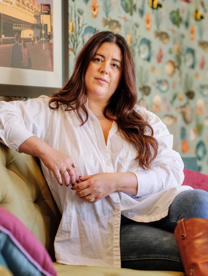 A woman in a white blouse sits on a couch indoors, looking toward the camera with a framed painting and patterned wallpaper in the background.