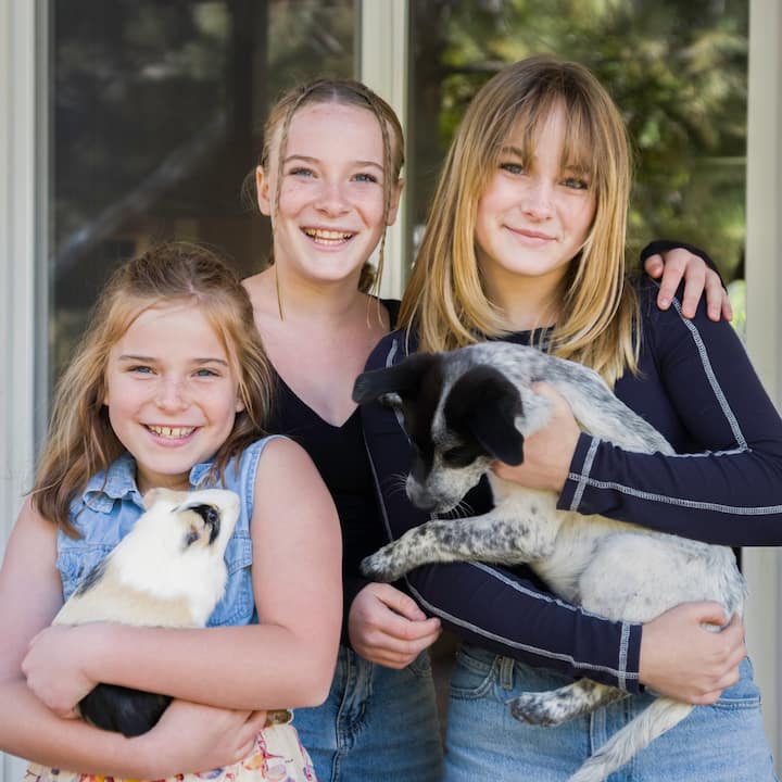 Three girls smiling together outdoors, with one holding a small animal and another holding a dog.