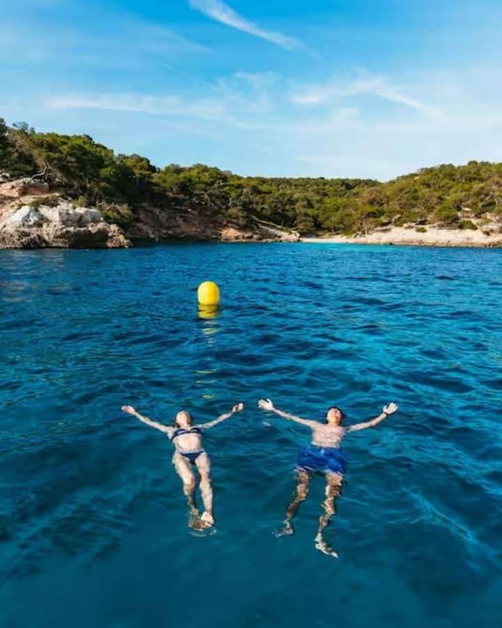 Una pareja flotando en un mar turquesa cerca de una costa rocosa y arbolada. La imagen representa las estancias tranquilas para ver el eclipse en las islas Baleares.