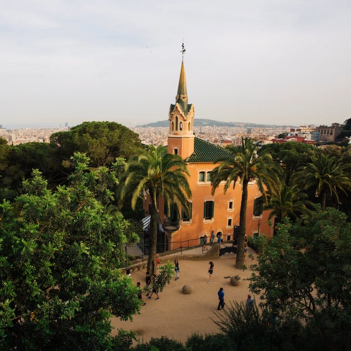 Vista desde la ladera de una colina de Barcelona de un edificio de Gaudí con agujas y una plaza, con la ciudad extendiéndose al fondo.