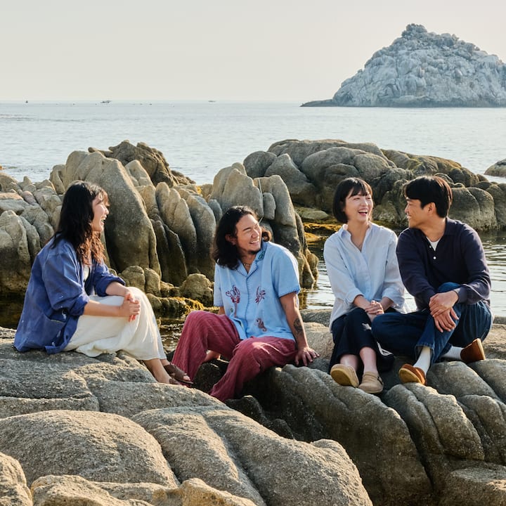 바닷가 바위 위에서 네 명이 편안하게 앉아 웃고 있는 장면, 함께 살아가는 듯한 여행의 여유
