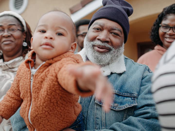 A man holds a baby outdoors while surrounded by family members smiling together in front of a home.