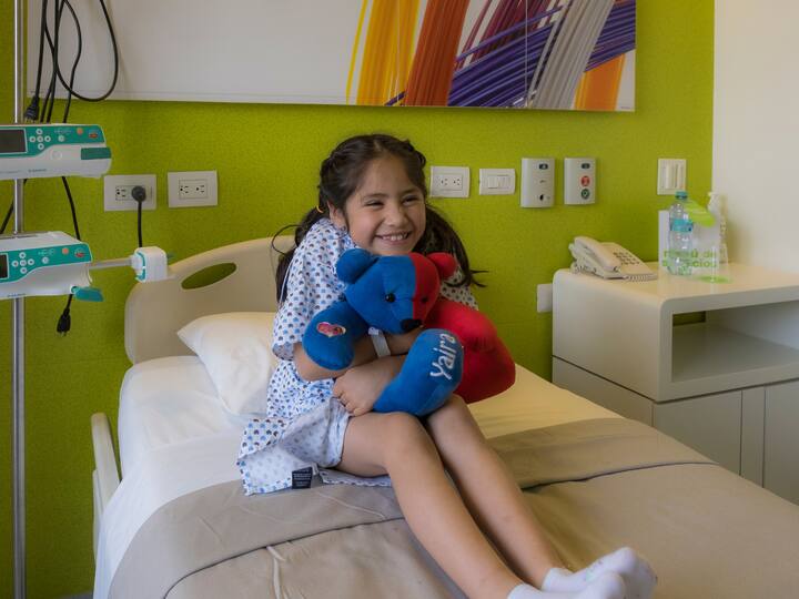 Seorang gadis muda dalam gaun hospital duduk di atas katil hospital sambil tersenyum dan memeluk patung haiwan.