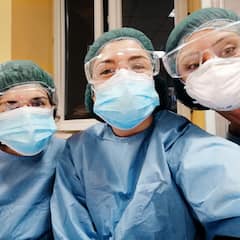 Dalawang healthcare worker na nakasuot ng surgical gown, mask, at cap na nakangiti para sa litrato.