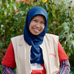 Isang babaeng nakangiti at nakasuot ng hijab at volunteer vest na nasa hardin sa labas.
