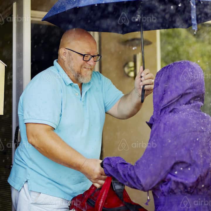 A man holds an umbrella over a person in a purple raincoat in the rain outside a home.