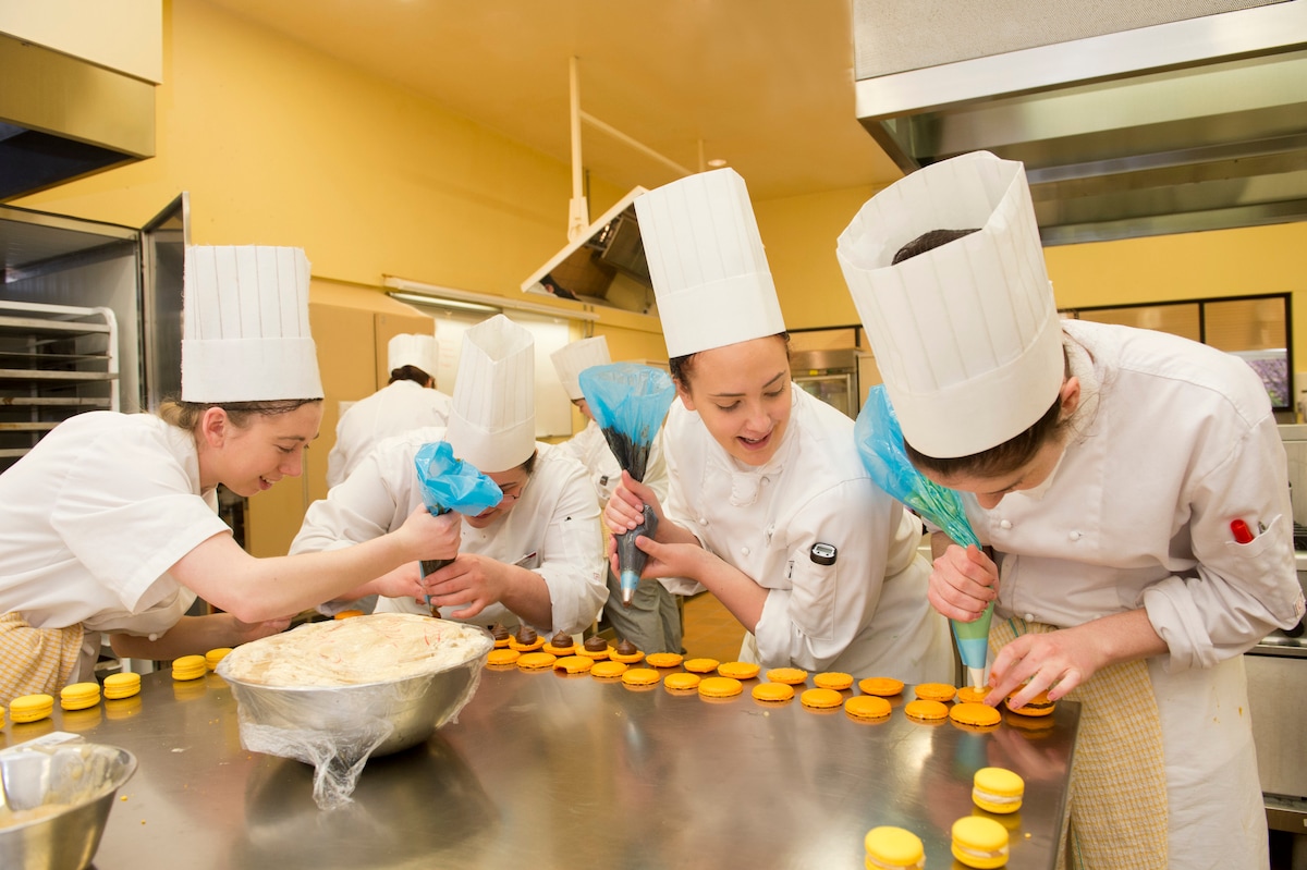 Guests in chef hats piping filling into rows of bright yellow macarons during a Paris baking class.