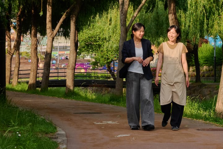 Suncheon host strolling with a guest on a shaded park path beside the river