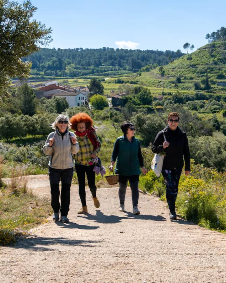 Un grupo de amigos con cestas caminan por un sendero rural entre sembrados en terrazas verdes a las afueras de Barcelona, bajo un cielo azul claro.