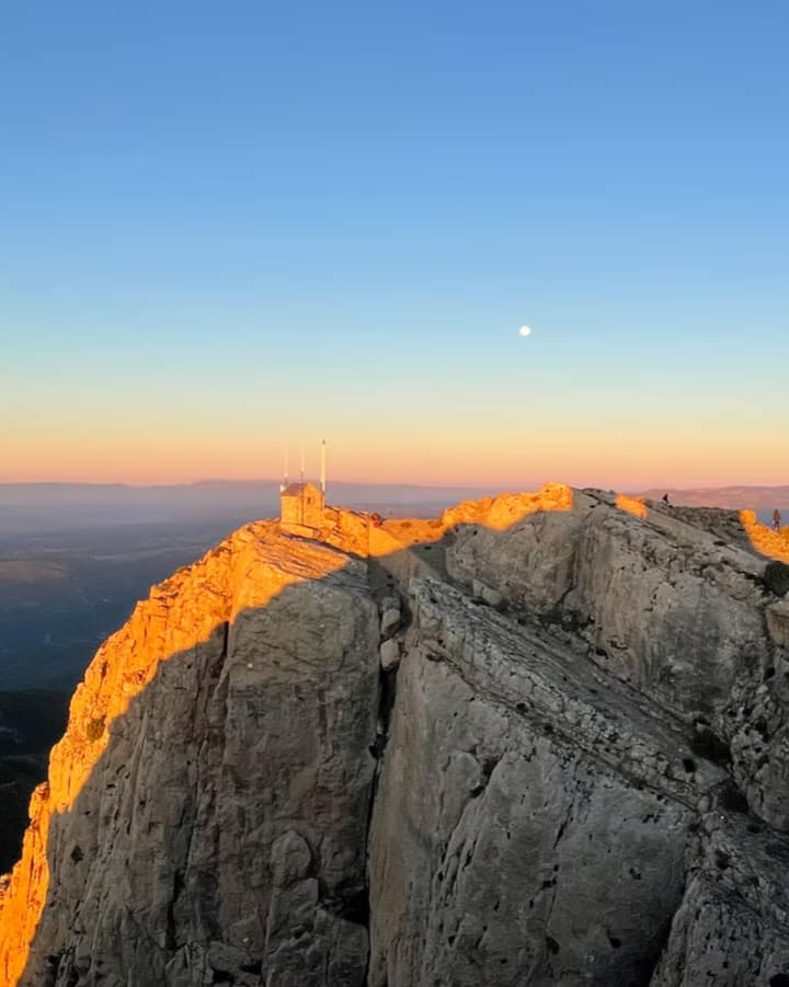 La luz dorada del atardecer ilumina una montaña valenciana, con vistas a valles y al cielo con la luna a lo lejos.