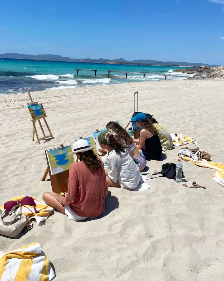 Un grupo de amigas sentadas en la arena de una plaza ibicenca pintan el mar; la imagen promociona las estancias en Airbnb para ver el eclipse en las islas Baleares.