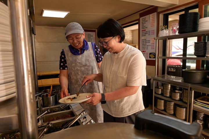 Host in Seongbuk-dong serving food in a cozy kitchen, smiling with a guest by the buffet counter