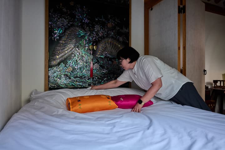 Seongbuk-dong host smoothing a neatly made bed with colorful silk pillows in a traditional room