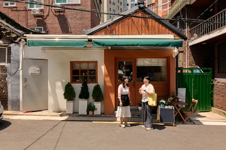 Two people talk in front of a cozy Seongbuk-dong house with potted plants and a café table