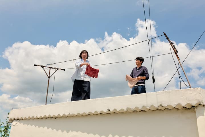 Two Goseong hosts smiling on a sunny rooftop, shaking out red and beige cushions under blue sky.